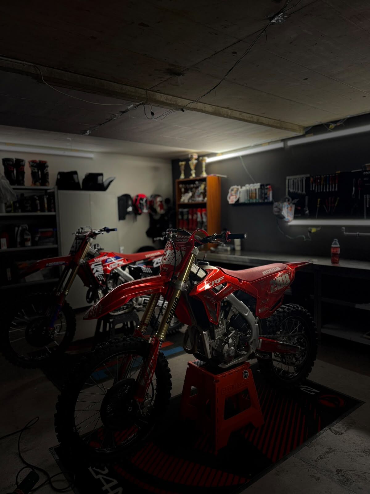 Le garage de Luks la nuit : deux Honda CRF rouges, trophées, casques et outillage au mur.